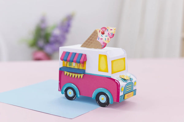 A colorful handmade paper toy ice cream truck parked on a light blue card on a pink surface. The toy features a white cab, a pink side, yellow windows, blue wheels, and a small paper cone on top.