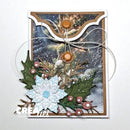 A handmade Christmas greeting card featuring a snowy winter scene with pine trees, holly leaves, berries, a white snowflake embellishment, and two brown buttons tied with white string. The card is photographed against a plain white background.