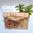A handmade decorative card or small box with layered paper elements, floral cutouts, twine bow, and a small potted green plant behind it, photographed against a plain light background. Large overlaid text reads 'Made by Corry for CREA.