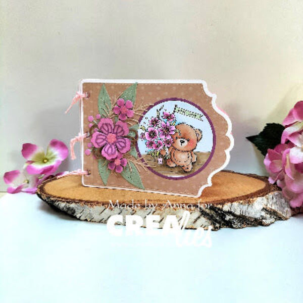 A handmade scrapbook card featuring a cute teddy bear illustration, pink flowers, and green leaves on a kraft paper background, displayed on a rustic wood slice with pink flowers in the background.