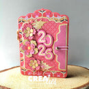 A handmade pink scrapbook journal with 3D layered paper flowers, scalloped edges, and the year '2026' in raised pink paper. The journal is displayed on a rustic wood slice against a neutral gray background.