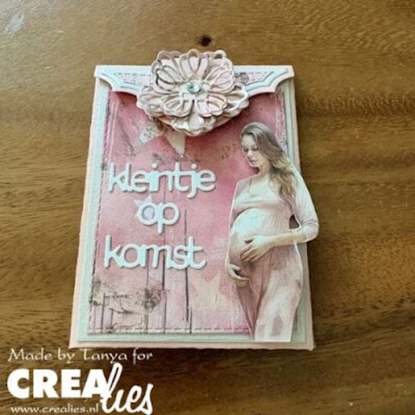 A handmade greeting card or decorative plaque on a wooden surface, featuring the Dutch text 'kleintje op komst' in white script over a pink distressed background, with a 3D flower embellishment at the top and a photo of a pregnant woman in a light pink dress. A watermark at the bottom left reads 'Made by Tanya for CreaLies'.