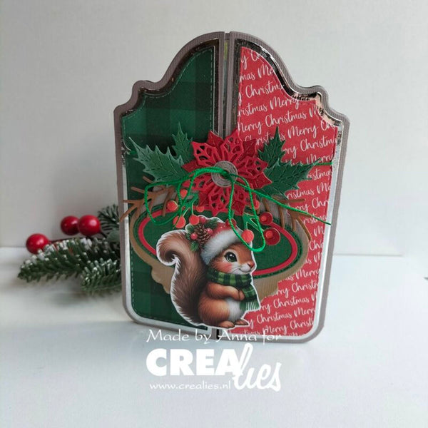 Handcrafted Christmas card or decorative sign shaped like a gift box, featuring a green plaid panel, red script background, holly leaves, berries, and a squirrel illustration with a Santa hat. A small pine branch with red berries sits beside it on a white surface against a light gray background.