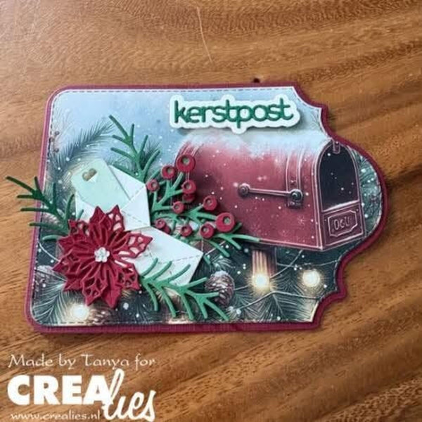 A handmade Christmas-themed paper cutout plaque featuring a red mailbox, green pine branches, red berries, a red paper flower, and the word 'kerstpost' on a snowy background, displayed on a wooden surface.