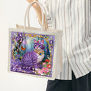 A person wearing a white and gray striped shirt holds a clear tote bag with a colorful printed illustration of a purple cat in a floral garden. The tote bag has white handles and a white fabric base.