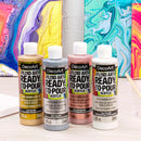 Four bottles of DecoArt Fluid Art Ready-to-Pour Acrylic paint in gold, silver, pink, and white, displayed in front of a colorful abstract canvas on a light wood surface.
