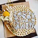 A set of silver metal floral and botanical die-cut stickers arranged on a round woven straw placemat, with dried flowers and a gold binder clip on a brown paper sheet beside it.