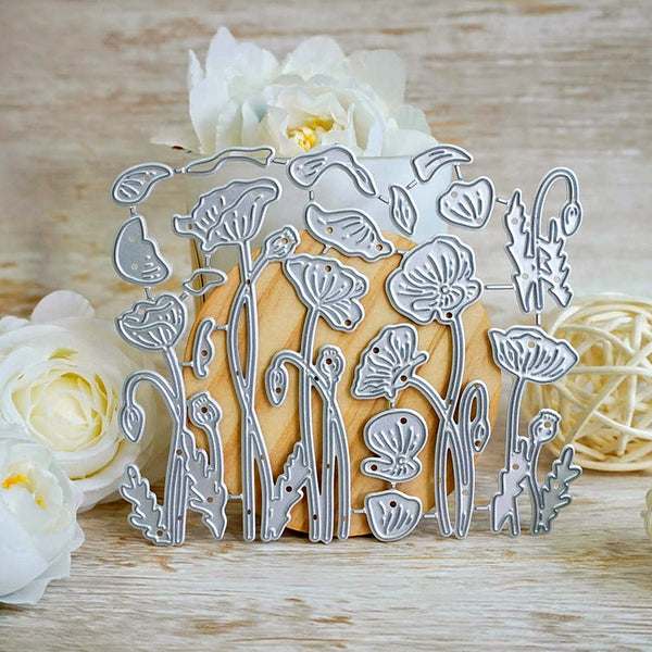 A silver metal die-cut floral design sheet featuring poppy and wildflower motifs, displayed on a wooden block with white flowers and a woven ball prop in the background.