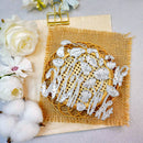 A flat-lay of a white laser-cut metal floral and botanical design sheet placed on a circular wooden coaster over a square piece of tan burlap fabric, styled with white flowers and cotton bolls on a light gray background.