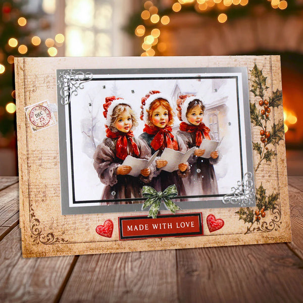 A decorative handmade Christmas greeting card displayed on a wooden surface with a blurred festive background. The card features a rustic beige border with ornate scrollwork, a central photo of three young girls in winter clothing holding sheet music, red hearts, a green bow, and a red banner that reads 'MADE WITH LOVE'.