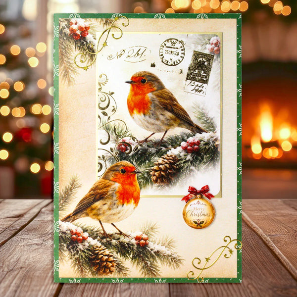 A rectangular Christmas greeting card or art print featuring two robin birds perched on snowy pine branches with vintage postage stamps and decorative flourishes. The card has a green border and is displayed upright on a wooden surface with a blurred warm holiday background.