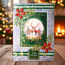 A festive Christmas greeting card displayed on a wooden table with a blurred warm holiday background. The card features a snowy globe illustration with two red deer, red poinsettias, green holly leaves, and a gold 'Warm Wishes' label.