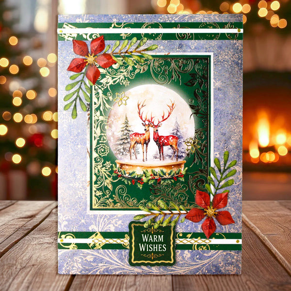 A festive Christmas greeting card displayed on a wooden table with a blurred warm holiday background. The card features a snowy globe illustration with two red deer, red poinsettias, green holly leaves, and a gold 'Warm Wishes' label.