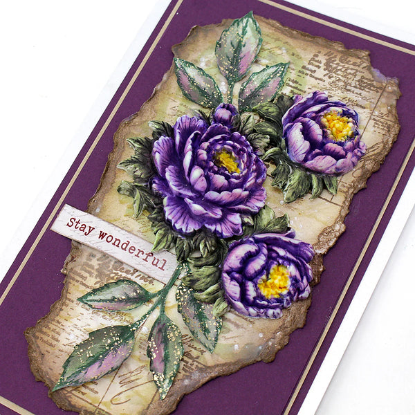 A close-up of a handmade greeting card featuring three purple peony flowers with yellow centers, green leaves, and a distressed beige paper background. The card has a deep purple border and a small white banner that reads 'stay wonderful'.