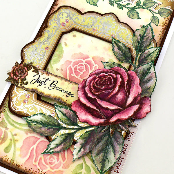 A close-up of a decorative handmade card featuring a large layered rose with green leaves, a scalloped ornate frame, floral background print, and a banner reading “Just Because.”.