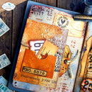 A rustic mixed-media paper collage journal page featuring vintage-style elements including ticket stubs, postmarks, stamps, and distressed paper textures in orange, beige, brown, and cream tones, arranged on a dark blue tray over a wooden surface.