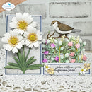 A handmade layered card featuring three white daisies with yellow centers, a small brown-and-white bird perched above a cluster of pink, purple, and white wildflowers, and the printed phrase 'Where wildflowers grow, happiness follows.' The card is displayed on a white lace surface against a light gray brick background, with a small hand visible in the top-left corner.