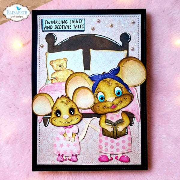 A handmade greeting card featuring two cute cartoon mice in bed, with one reading a book and the other standing nearby. The card has a black border and is displayed on a pink textured surface with warm bokeh lights in the background.