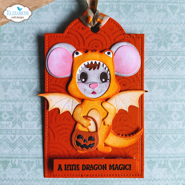 A handmade Halloween ornament featuring a layered orange dragon mouse with pink ears, a pumpkin bucket, and the text 'A LITTLE DRAGON MAGIC!' on a light blue patterned background. The brand logo 'Elizabeth craft designs' is visible in the top left corner.