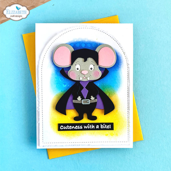 A handmade greeting card featuring a cute vampire mouse character with pink ears, a black cape, and a purple collar, set against a blue and yellow background. The card has the text 'Cuteness with a bite!' and is shown with a yellow envelope on a light blue surface.