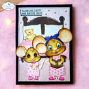 A handmade greeting card featuring two cute cartoon mice in bed, with one reading a book and the other standing nearby. The card has a black border and is displayed on a pink textured surface with warm bokeh lights in the background.