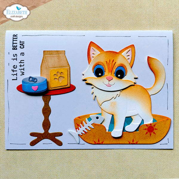 A handmade layered paper craft card featuring a cute orange and white cat sitting in an orange bowl, next to a small table with a blue can and a bag of cat food. The card has the text 'Life is BETTER with a CAT' and is displayed on a beige textured fabric background.