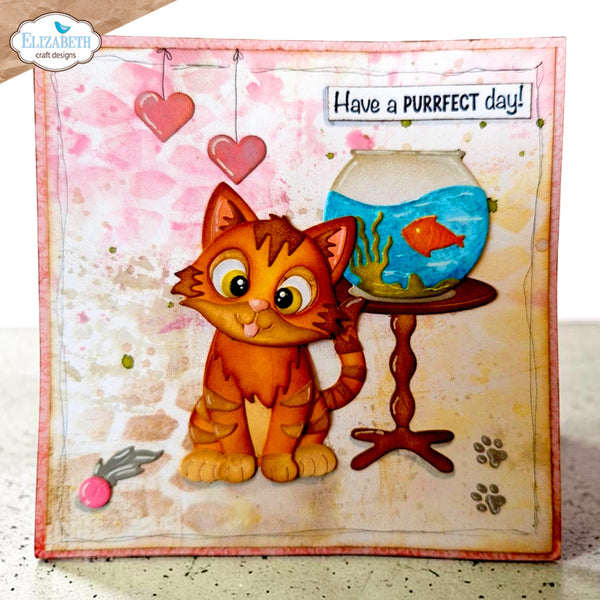 A handmade greeting card featuring a cute orange tabby cartoon cat next to a fishbowl with a goldfish, with the text 'Have a PURRFT day!' on a watercolor-style pink and beige background. The card is shown standing upright on a speckled countertop.