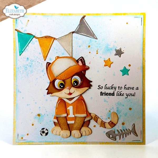 A handmade square greeting card featuring a cute cartoon cat dressed in an orange soccer jersey and cap, with colorful bunting, stars, a fish skeleton, and the text 'So lucky to have a friend like you!'. The card has a white center, a yellow border, and a blue watercolor splash background.