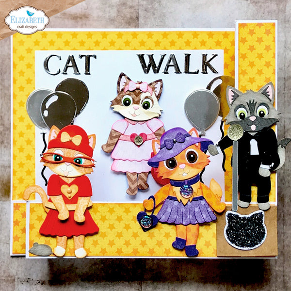 A handmade craft scrapbook card featuring three cute cartoon cats in different outfits under the text 'CAT WALK'. The card has a yellow star-patterned border, a white center, and is displayed on a neutral textured surface.