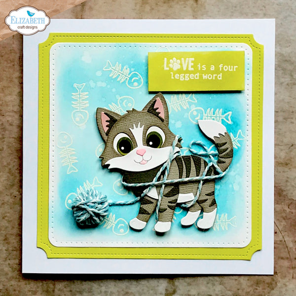 A handmade greeting card featuring a 3D papercat holding a ball of yarn, set against a light blue watercolor background with fish skeleton motifs. The card has lime green borders, a white cardstock base, and a yellow sticker that reads 'LOVE is a four legged word'.