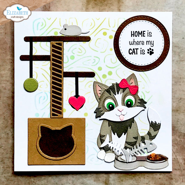 A square handmade greeting card featuring a cartoon tabby cat with green eyes and a pink bow, sitting beside a cat tree, fish, food bowl, and a circular sign that reads 'HOME is where my CAT is'. The card is photographed on a textured beige surface, with an 'Elizabeth craft designs' watermark in the top-left corner.