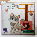 A square handmade greeting card or art print featuring a cute gray tabby cat, a wooden cat tree, a red cat house, a green double bowl, a purple yarn ball, and the text 'Life is BETTER with a CAT'. The card has a white background with subtle lavender floral patterns, a purple border, and is shown standing on a white surface.
