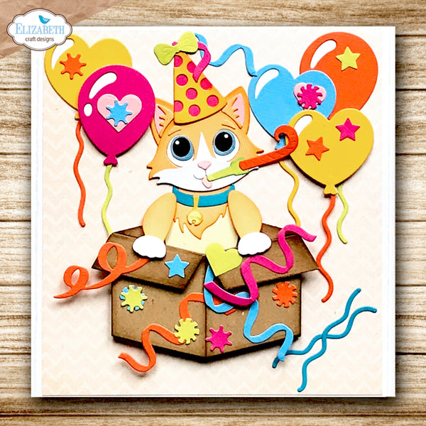 A square craft design or card featuring a cartoon kitten sitting in an open box with colorful balloons, party hat, and streamers, shown on a light wood background. The top-left corner includes a watermark reading Elizabeth craft designs.