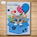 A handmade birthday card for kittens, featuring a gray kitten popping out of a blue box with balloons, stars, and the text 'Another birthday? You gotta be kitten me!'. The card is displayed flat on a light wood background.