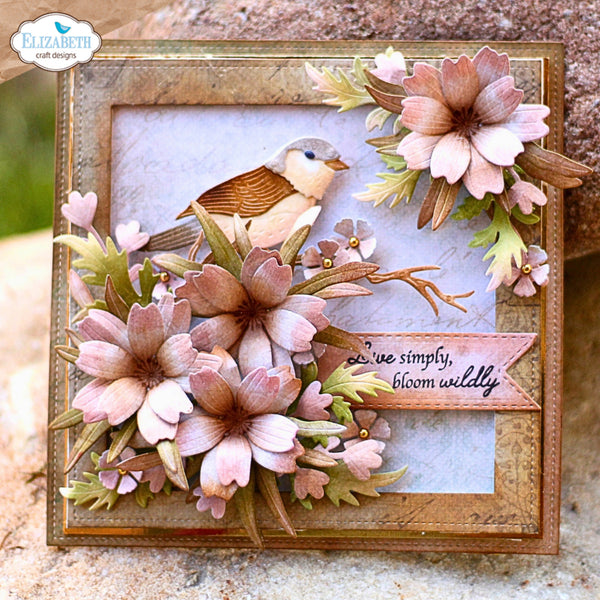 A square handmade greeting card featuring a small bird perched among layered pink and beige flowers with green leaves, set in a decorative gold-toned frame. A ribbon banner reads, 'we've simply, bloom wildy.