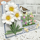 A handmade layered card or decorative plaque featuring three large white daisy flowers with yellow centers, green leaves, a small brown-and-white bird, and a small floral arrangement with the phrase 'Where wildflowers grow, happiness follows.' The product is photographed on a white textured surface against a white brick wall, with a visible hand at the top left and a 'Elizabeth craft designs' logo in the upper left corner.
