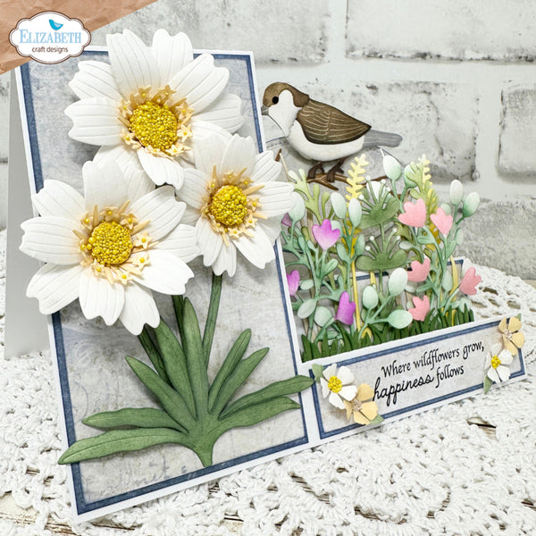 A handmade layered card or decorative plaque featuring three large white daisy flowers with yellow centers, green leaves, a small brown-and-white bird, and a small floral arrangement with the phrase 'Where wildflowers grow, happiness follows.' The product is photographed on a white textured surface against a white brick wall, with a visible hand at the top left and a 'Elizabeth craft designs' logo in the upper left corner.