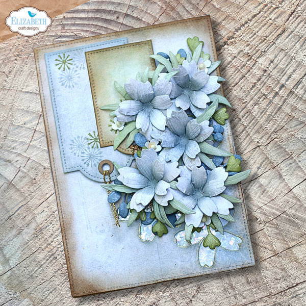 A handmade scrapbook card featuring layered paper flowers in pale blue, white, and sage green, with a small vintage key charm detail, displayed on a rustic wood slice background. The brand logo 'Elizabeth craft designs' is visible in the top-left corner.