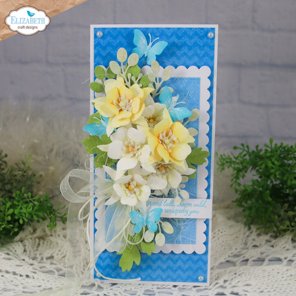 A handmade layered card with blue chevron and solid backgrounds, decorated with yellow and white paper flowers, green leaves, blue butterflies, and a sheer white ribbon bow. The card is displayed on a white lace runner with small green plants in the background.