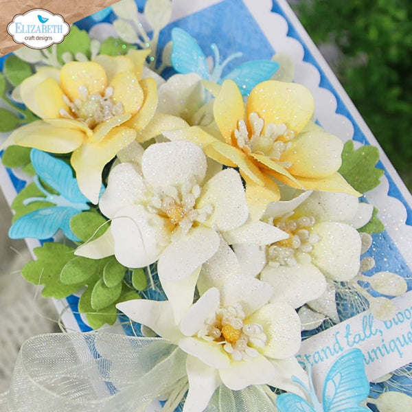 A close-up of decorative artificial flowers arranged on a blue gift box. The flowers include pale yellow blossoms, white blossoms with glittery centers, green leaves, and light blue butterfly accents.
