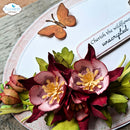 A close-up of a handmade floral embellishment or scrapbook decoration featuring layered pink and burgundy flowers with yellow centers, green leaves, and a small wooden butterfly on a white circular backing. A brand watermark in the top left reads 'Elizabeth craft designs', and part of a card says 'Cherish the wildflower unscripted'.