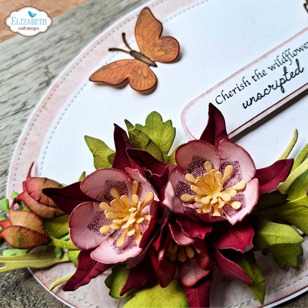 A close-up of a handmade floral embellishment or scrapbook decoration featuring layered pink and burgundy flowers with yellow centers, green leaves, and a small wooden butterfly on a white circular backing. A brand watermark in the top left reads 'Elizabeth craft designs', and part of a card says 'Cherish the wildflower unscripted'.