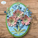 A 3D layered floral oval plaque featuring three textured paper flowers in shades of dusty pink and terracotta, accented with green and blue foliage on a light blue background, all mounted on an oval green frame. The plaque is displayed flat on a wooden surface.