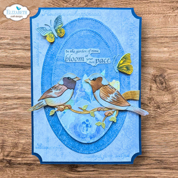 A handmade layered card featuring two 3D bird cutouts on a blue oval background with butterflies and the phrase 'in the garden of time bloom at your own pace.' The card is photographed flat on a wooden surface, with a 'Elizabeth craft designs' logo in the top-left corner.