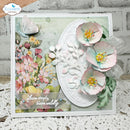 A handmade floral greeting card featuring three layered pastel pink and mint paper flowers, a white embossed oval center, and a watercolor-style floral panel on the left. The card is displayed on a white lace surface against a white brick wall background, with a 'Elizabeth craft designs' logo in the top-left corner.