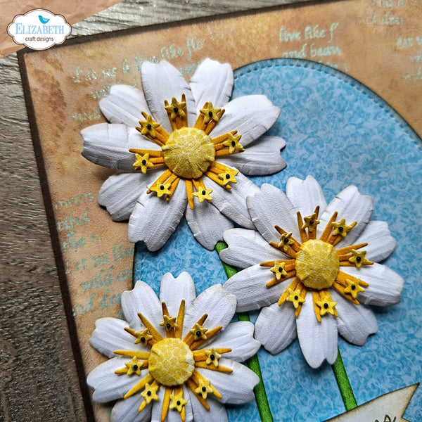 Three dimensional paper daisy embellishments with white textured petals, yellow centers, and yellow stamens, displayed on a blue patterned background with a brown textured card.