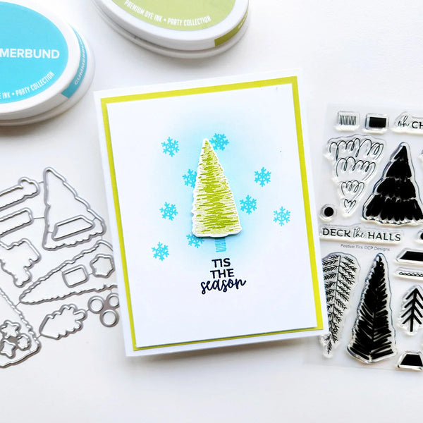 A handmade Christmas greeting card with a green pine tree and blue snowflakes, styled with crafting tools and ink pads on a white surface.