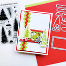 A handmade Christmas card with layered tree and banner designs, shown alongside clear stamp sheets and metal die cutters on red and white cardstock.