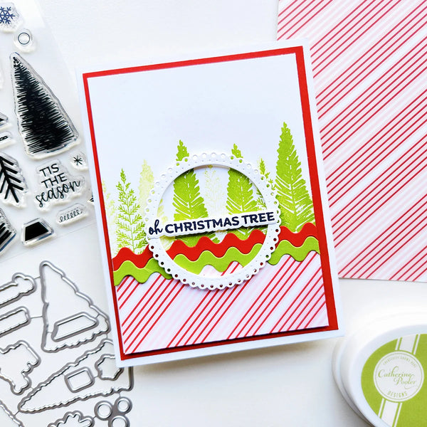 A handmade Christmas greeting card with layered red and white striped borders, green pine trees, and a central circular die-cut element reading 'Oh Christmas Tree,' shown on a white surface with crafting tools and patterned paper around it.