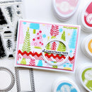 A handmade Christmas card featuring colorful stamped trees, snowflakes, stripes, and a die-cut circle with the text 'of CHRISTMAS TREE', surrounded by crafting tools and ink pads on a white background.